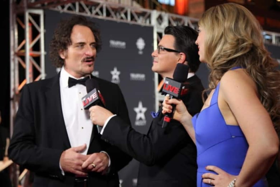 A dark-haired man in a tux talks to two reporters holding mics at a red carpet event.