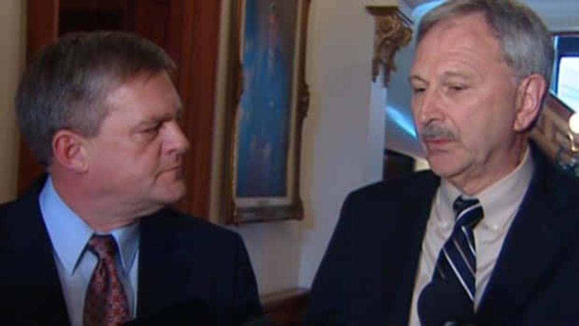 Two men in dark suits stand in front of a microphone.