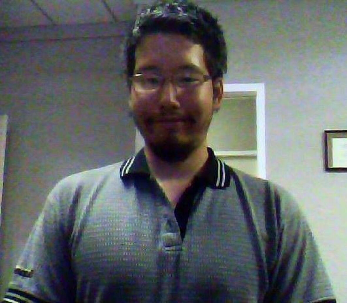 A grainy, dark image of a young man with glasses smiling faintly. He is wearing a grey polo.
