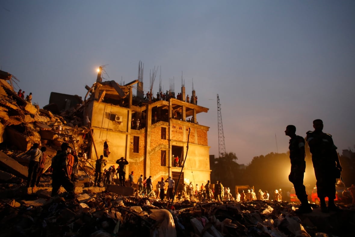 A building surrounded by rubble and people is lit up with floodlights against a darkening sky.
