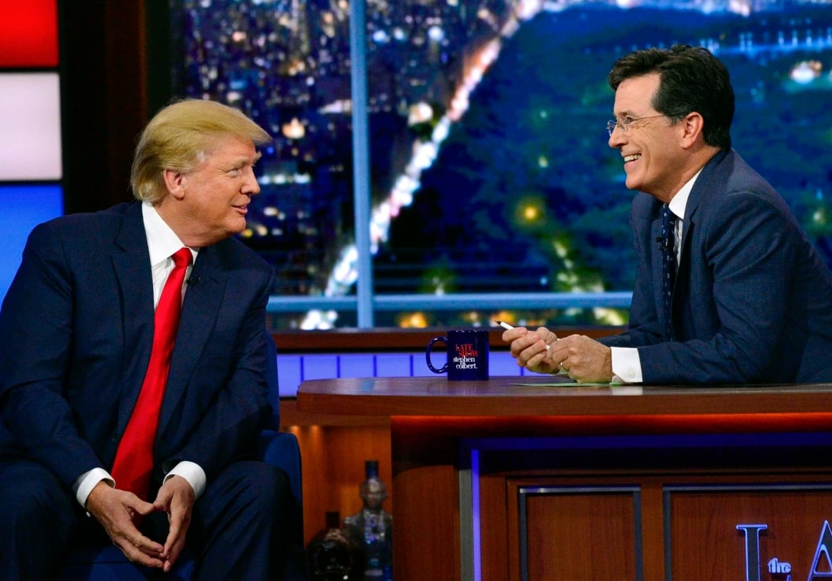A man with slicked back blond hair, wearing a dark suit, white shirt and red tie, sits on a chair facing another man, wearing a navy suit and tie and white shirt, who is sitting behind a desk and smiling.
