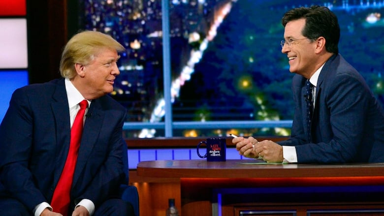 A man with slicked back blond hair, wearing a dark suit, white shirt and red tie, sits on a chair facing another man, wearing a navy suit and tie and white shirt, who is sitting behind a desk and smiling.