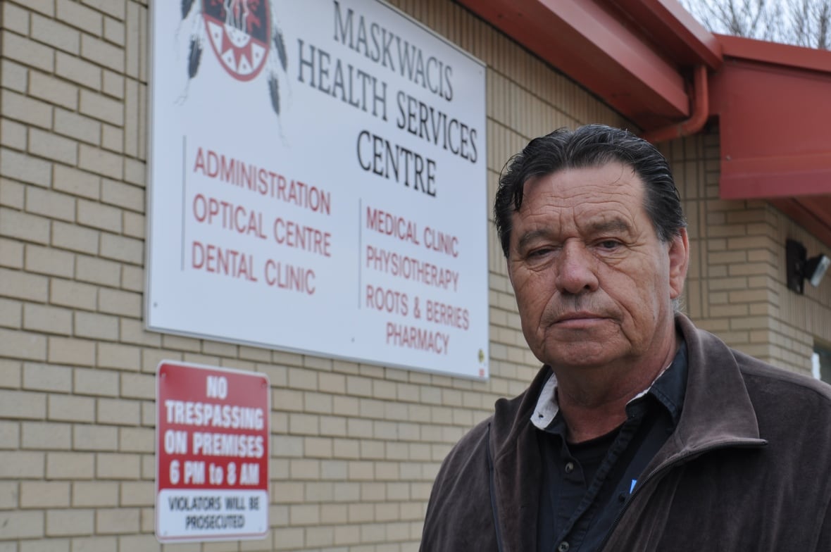 A man in a dark leather jacket with dark hair stands in front of a light brick building with a sign reading: Maskwacis health centre. 
