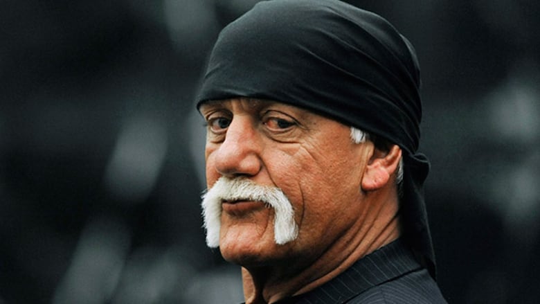 A man with a white handlebar mustache and a black cap around his head waits in a courtroom.