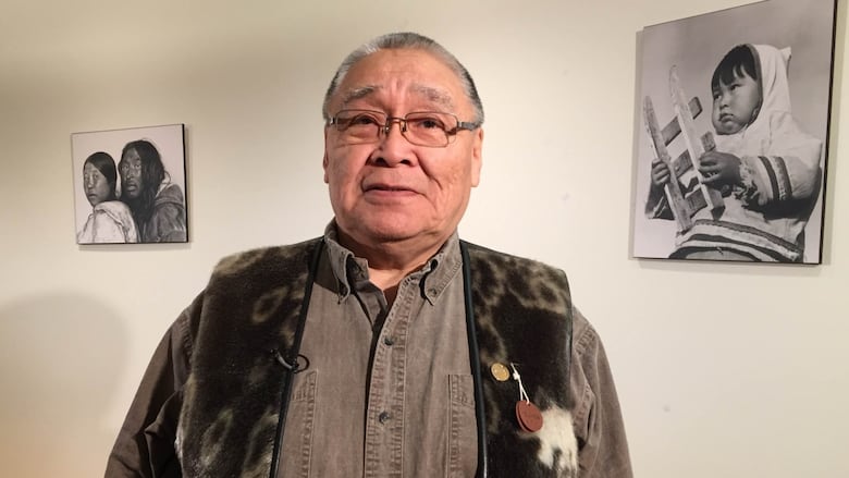 An older man in a sealskin vest stands in front of a wall with old photographs.