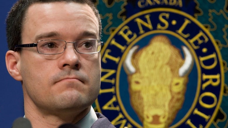 A man with very short, dark hair and glasses purses his lips, while speaking into a microphone in front of an RCMP emblem. 
