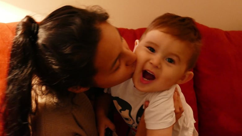 A teenage girl with long black hair in a ponytail kisses a baby boy, who is smiling at the camera.