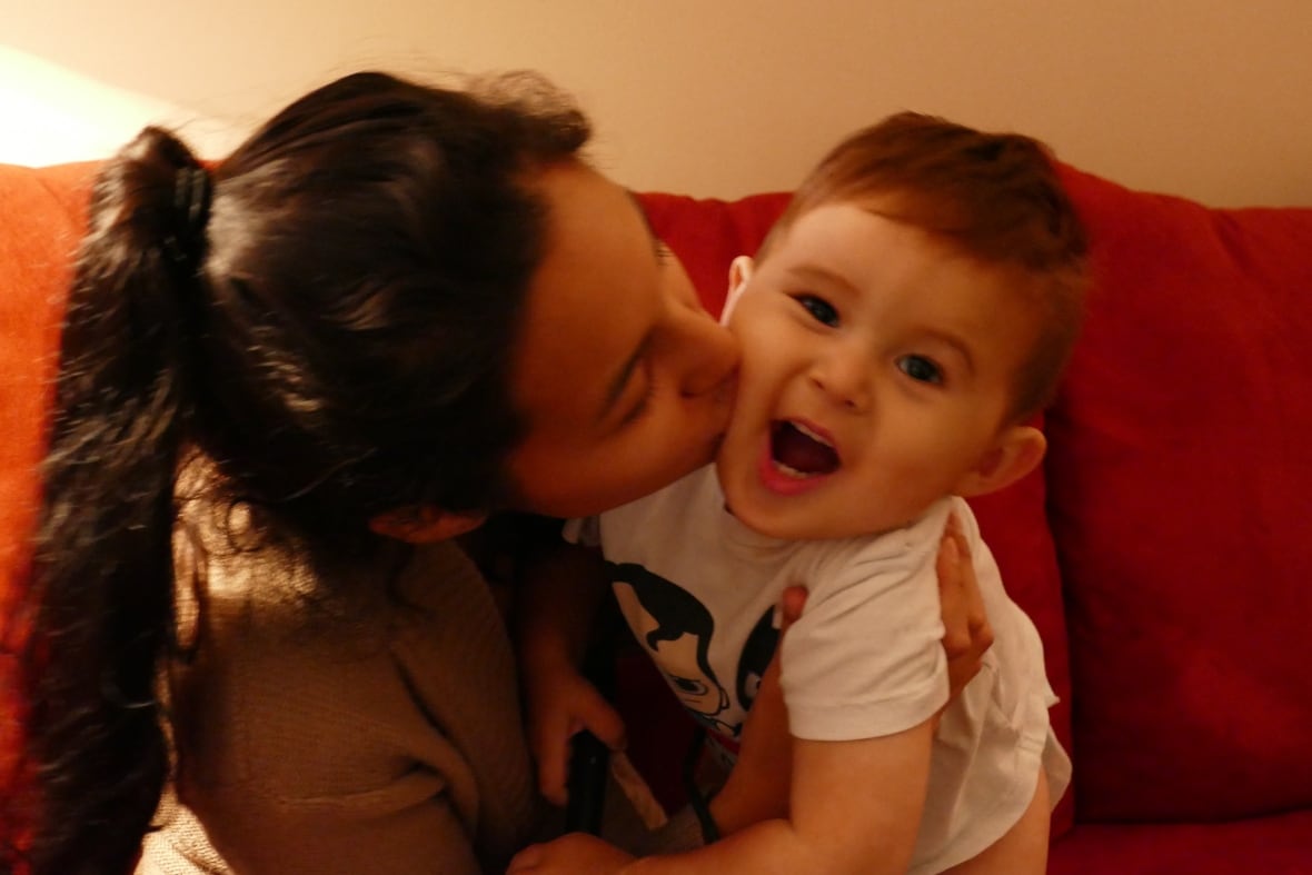 A teenage girl with long black hair in a ponytail kisses a baby boy, who is smiling at the camera.