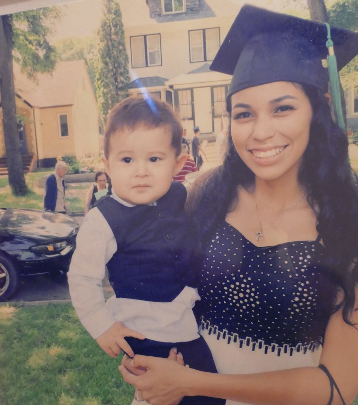 A teenage girl, wearing a white and black dress and a black graduation cap, holds a toddler while smiling into the camera.