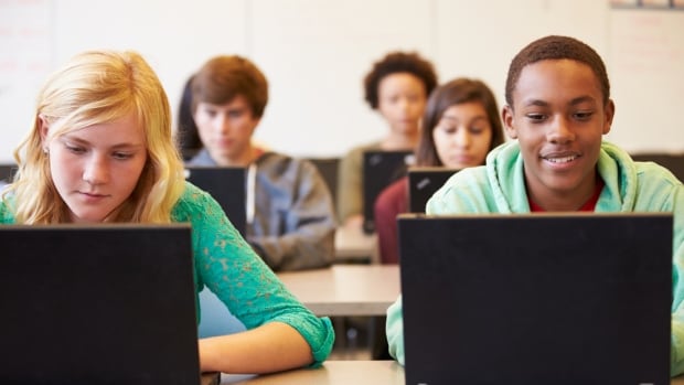 Teenage students in a classroom are seen sitting in rows, looking down at open laptop computers before each of them.