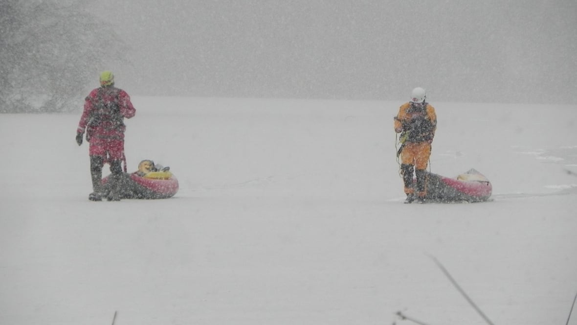 Two people in rescue gear pull sleds through blowing snow on a frozen lake.