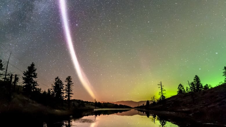 A purple streak of light in a starry night sky over a lake.