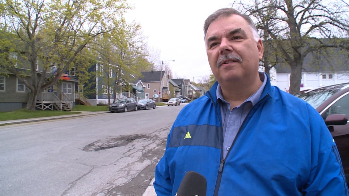 A man with a moustache and greying hair, and wearing a blue jacket, is standing on a sidewalk, with a paved road and houses visible in the background.