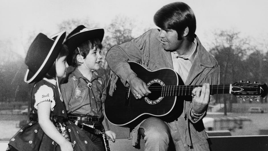 Campbell plays the guitar for two young fans, Renata Romoli and Francis Bradford, wearing cowboys hats in April 1970.