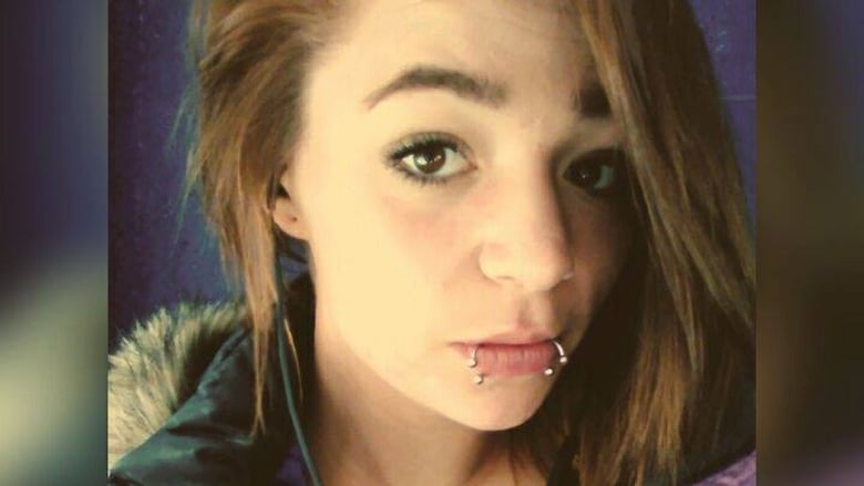 A portrait image of a young Caucasian woman with shoulder-length brown hair, brown eyes. She has two lip piercings, hoops with baubles. 