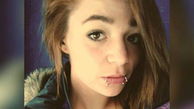 A portrait image of a young Caucasian woman with shoulder-length brown hair, brown eyes. She has two lip piercings, hoops with baubles. 