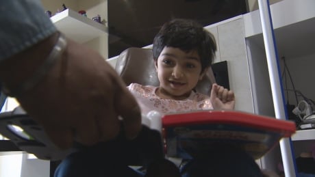 Amber Athwal sits in a chair in her home as her father encourages her to repeat the names of colours on a toy. Amber Athwal