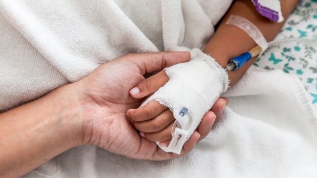 Alberta's pediatric hospitals are dealing with a wave of respiratory illnesses, including RSV, influenza and COVID-19. The number of severe cases is growing, driving hospital admissions up An adult holds a child's hand that has an IV in it. The hands are against a white blanket.