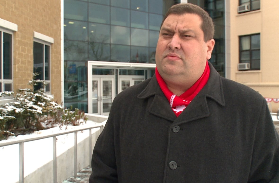 A man with a black coat and red scarf stands in front of Corner Brook city hall.