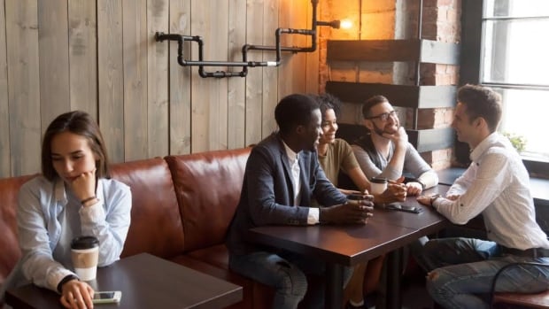 A woman, at left, looks down sadly as she sits alone at a table in a coffee shop. At a neighbouring table to the right, a smiling group of four people chat. 