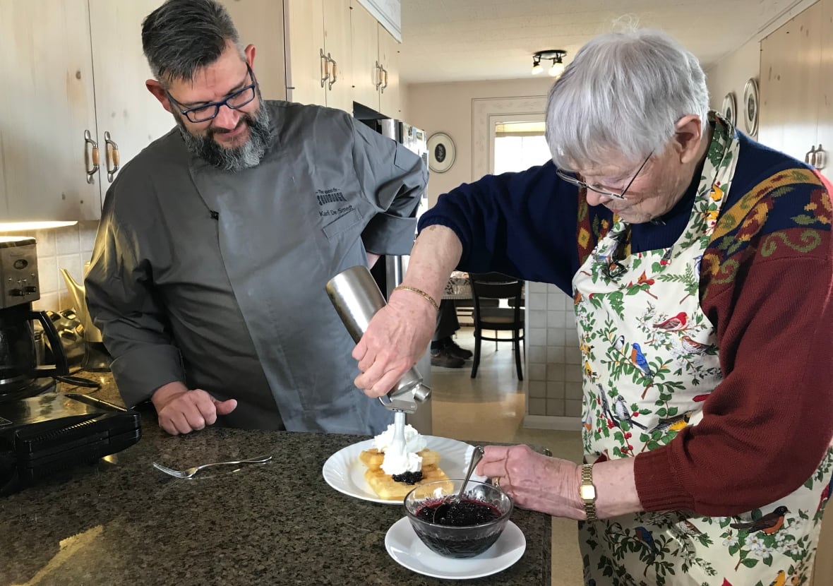 A woman stands in a kitchen and puts whipped cream on a pastry as a man looks on.