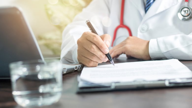 A stock image of a doctor, visible from the neck down, sitting at a desk, wearing whites and a stethoscope, filling in a form.