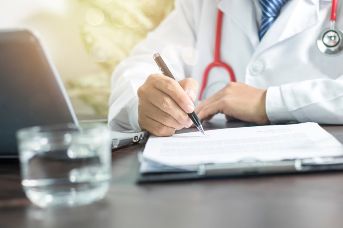 A stock image of a doctor, visible from the neck down, sitting at a desk, wearing whites and a stethoscope, filling in a form.