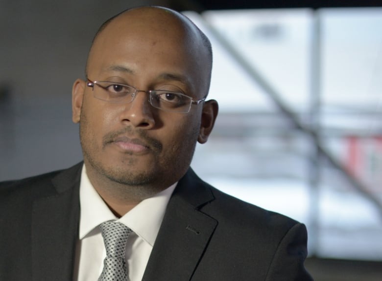 A man wearing glasses is shown in a dark suit.