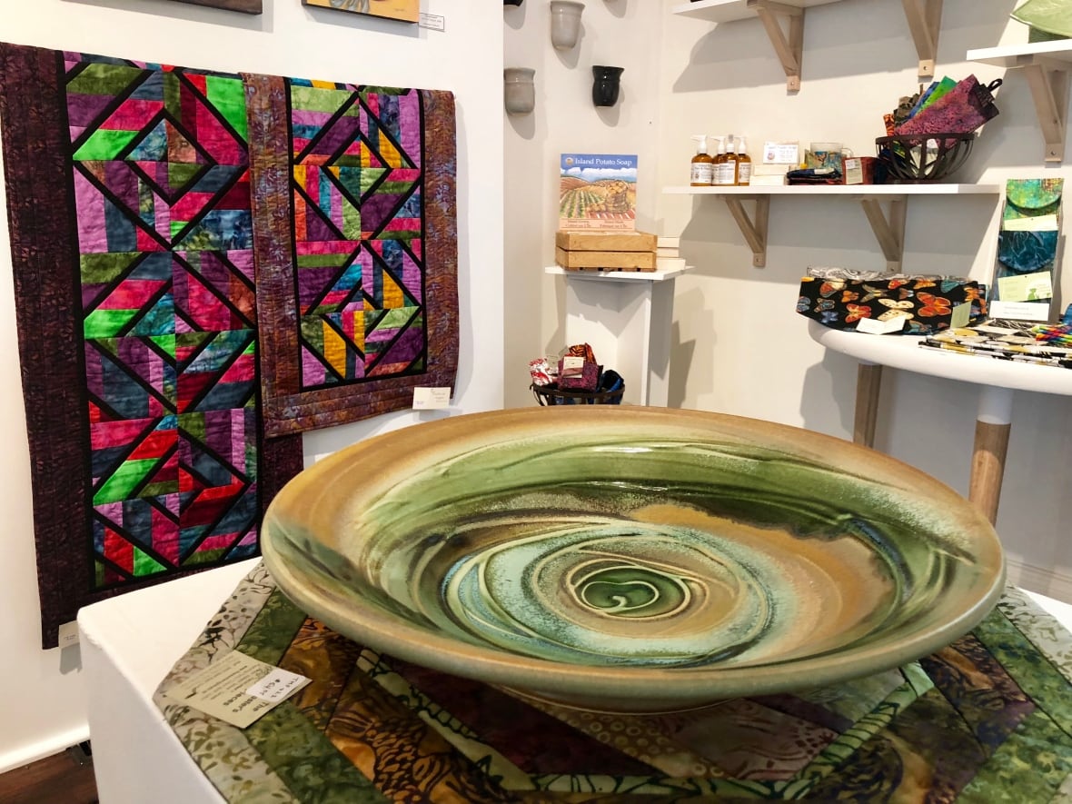 Some multi-coloured quilted wall hangings appear behind a large green and brown pottery bowl in a craft store.