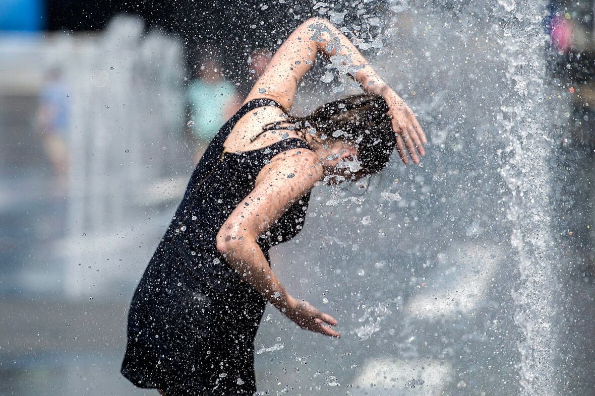 A woman in a black dress stands under water spouted from a fountain.