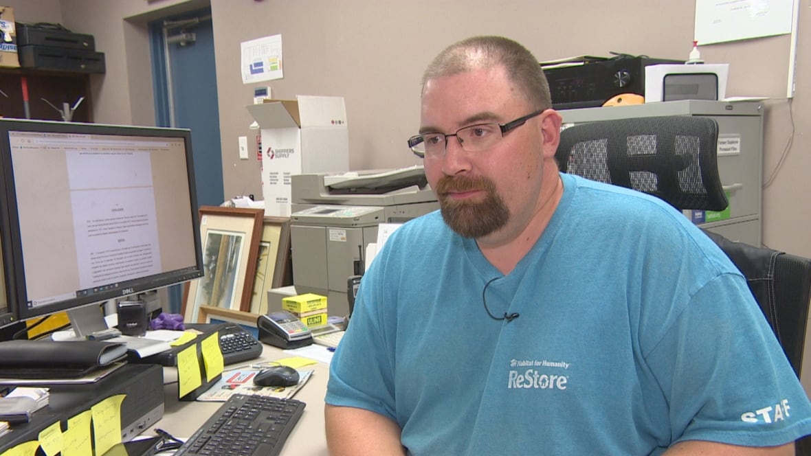 A man with a goatee and glasses and a blue shirt sits at a desk in front of a computer.