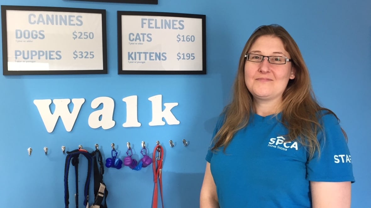 A woman with long hair and glasses stands in front of a blue wall.