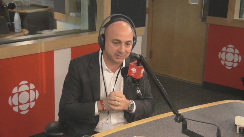 A man in a suit is wearing headphones and is speaking into a microphone with the CBC logo on it. 