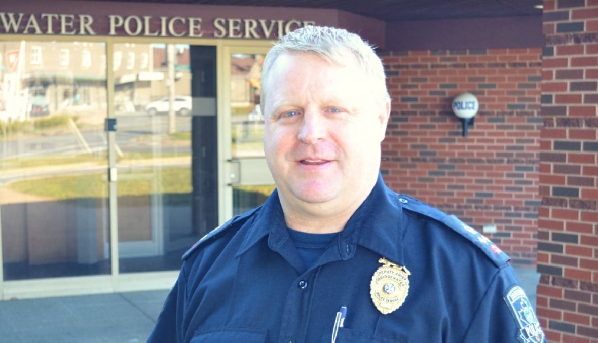 A white man with blonde hair wears a navy police uniform and stands outside a brick building