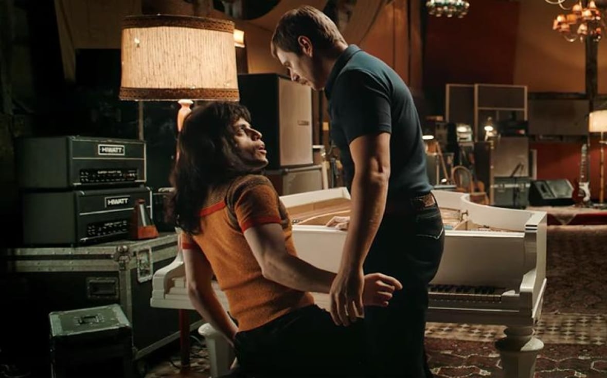 A man with long hair on the floor, looks at a second man standing directly to his right. The two closed the eyes and seem to be an intimate moment