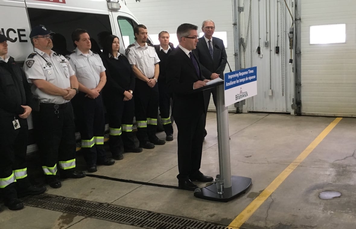 A man, Kris Austin, standing at a podium in front of a line of paramedics and another man, Blaine Higgs, in the corner.