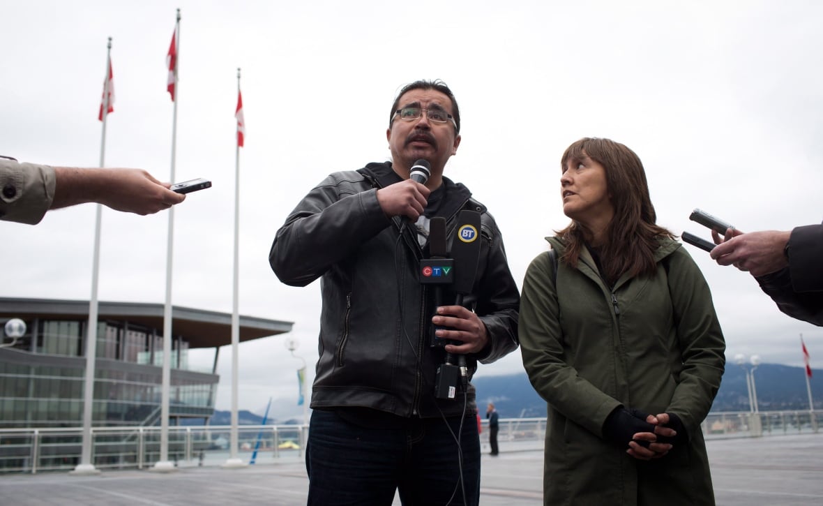 A man with a microphone with a woman beside him, by the Vancouver harbour front.