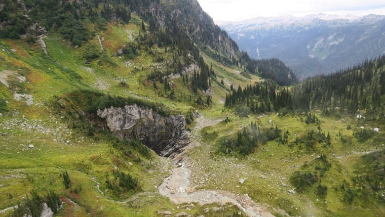 A wide shot of a cave in Wells Gray Provincial Park shows a pathway leading to a cave and a series of mountains in the background.