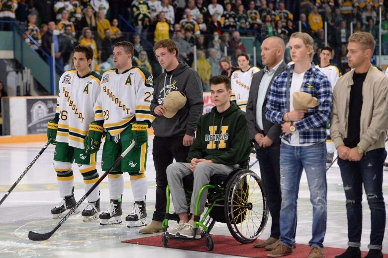 A group of men stand in a hockey rink, wearing shirts and jerseys reading Humboldt Broncos. The one in the centre sits on a wheelchair atop a red carpet.