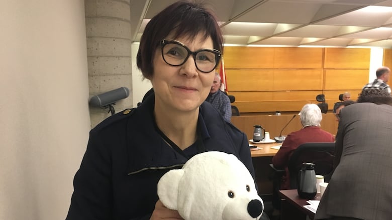 A woman with short hair and glasses holds a white teddy bear.