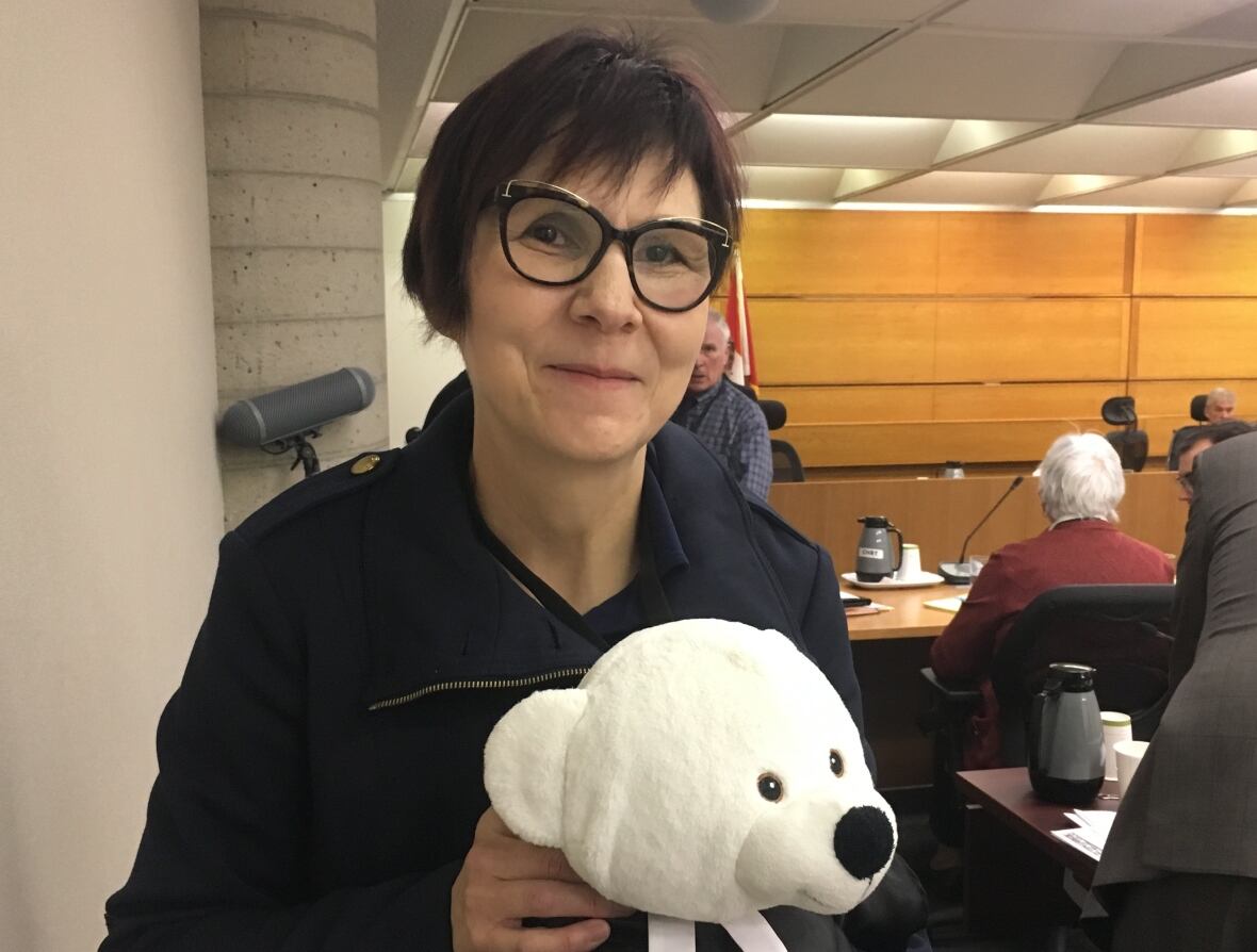 A woman with short hair and glasses holds a white teddy bear.