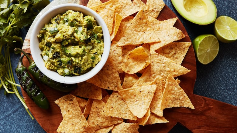 A bowl of guacamole on a wooden cutting board with tortilla chips on it. Grilled jalapeños, sliced lime and avocado sit next to it.
