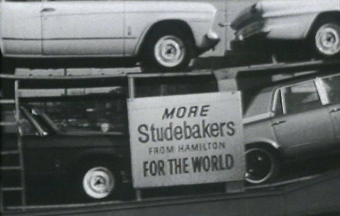 A black and white photo showing cars on a transport trailer with a sign on the trailer reading: "More Studebakers from Hamilton for the world."
