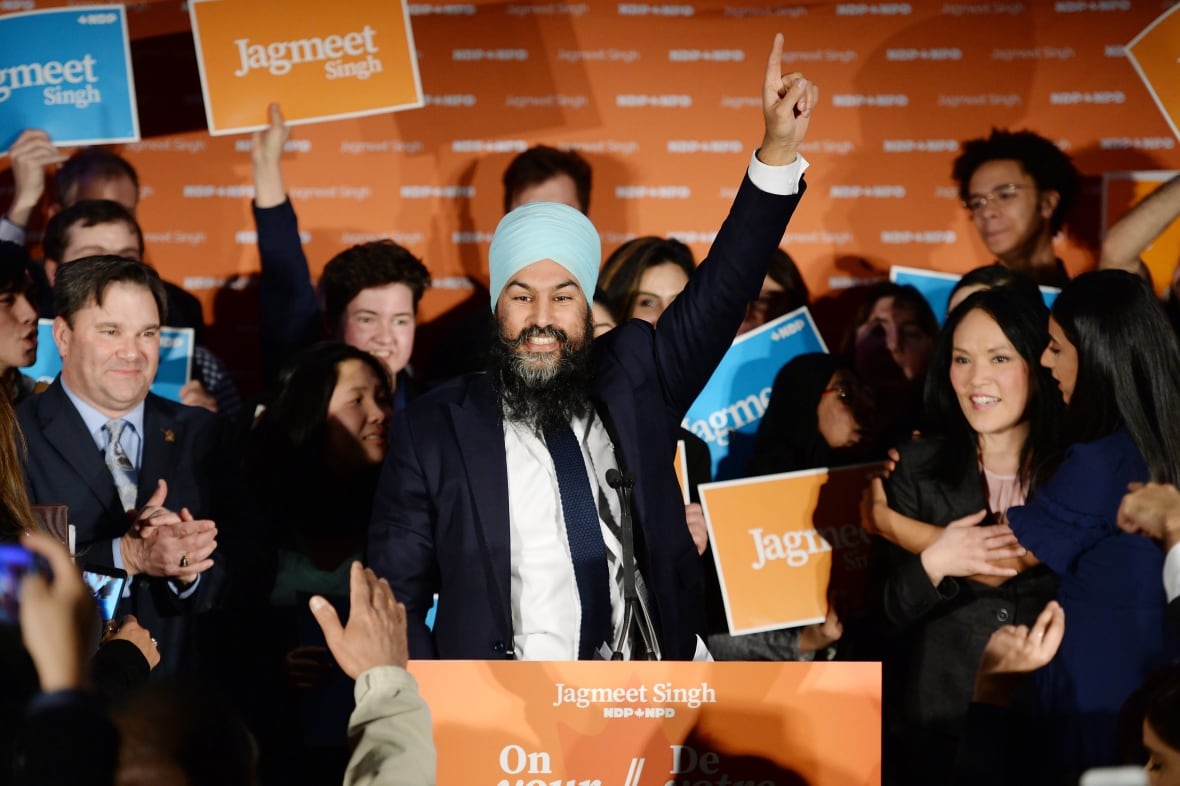 A man in a suit raises a hand with his index finger pointing up as a crowd applauds.
