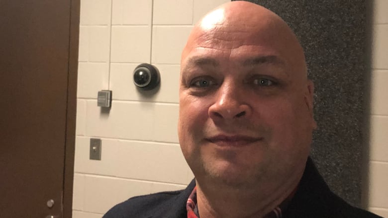 A bald man has a slight smile. A circular surveillance sits on the wall behind him.
