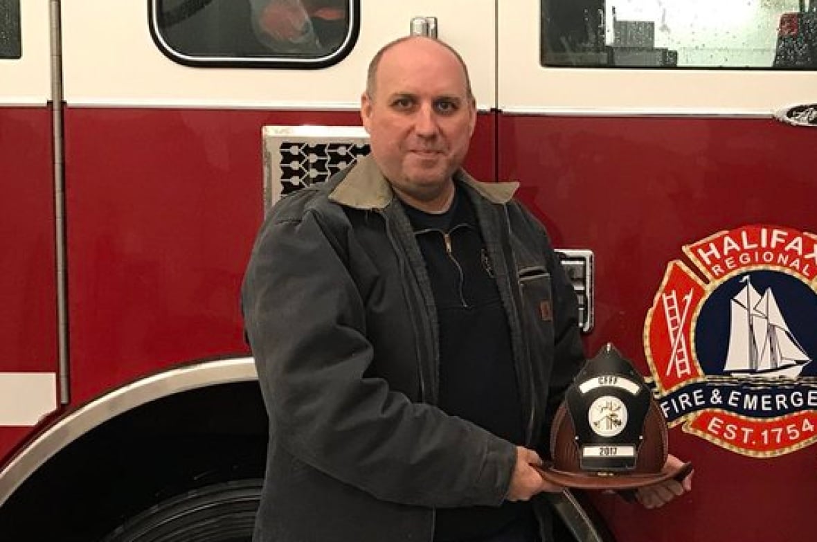 Two white men stand in front of a fire truck while holding fire fighter helmets