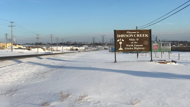 A wooden sign at the entrance to a snowy community reads, 'Welcome to Dawson Creek, Mile 0 of the World Famous Alaska Highway.'