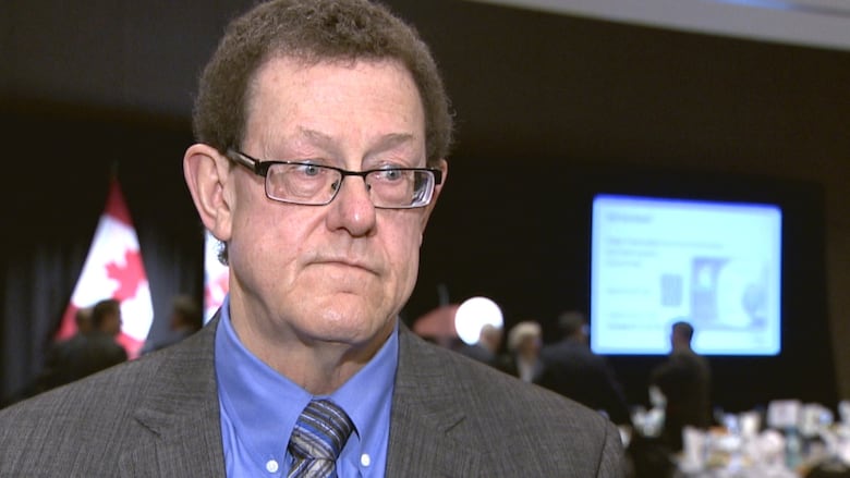 A man with brown hair looks just off camera with a serious expression. He is wearing glasses, a grey suit jacket, a blue dress shirt, and a blue and grey tie.