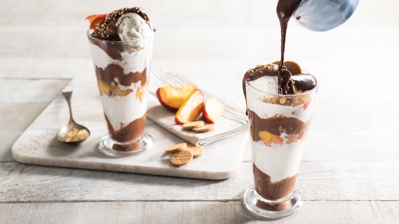 Hot fudge being poured from a small blue dish into a sundae in a tall glass with layers of vanilla ice cream, fudge and peaches. A marble tray with peach slices and another sundae sit behind it. It all sits on a grey wood surface.