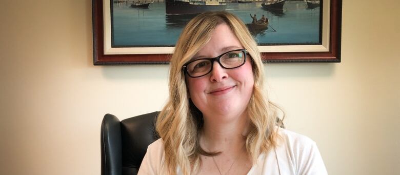 A woman with glasses sits at a desk and smiles.